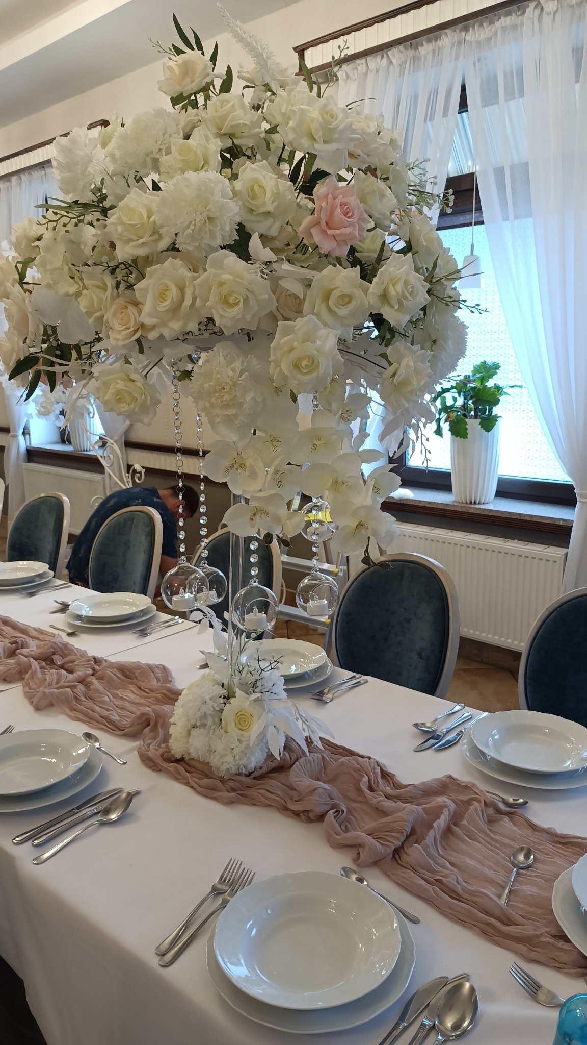 Uroczysta dekoracja stołu weselnego z wysoką kompozycją z białych róż i piwonii, szklane kule z tealightami zwisające z góry, pudrowy bieżnik na białym obrusie, elegancka zastawa.