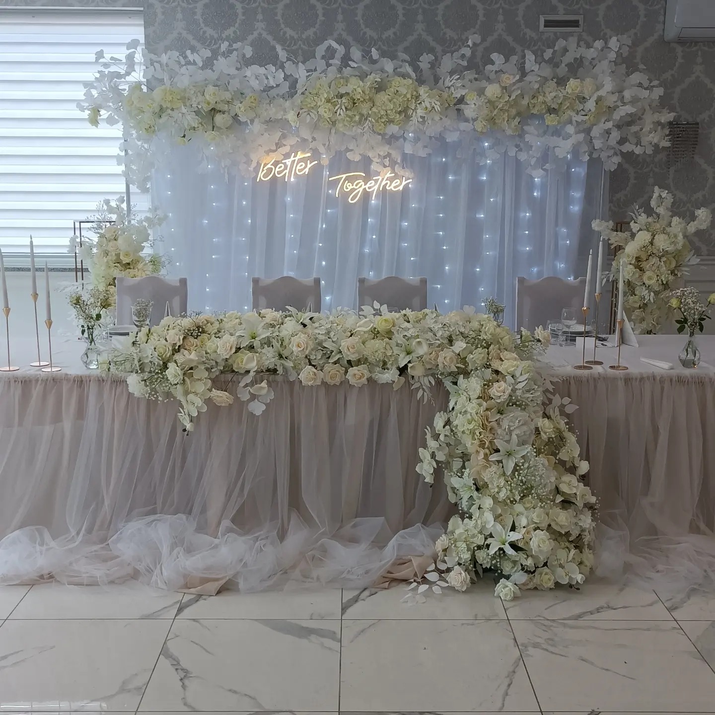 Elegancko udekorowany stół weselny z białymi kwiatami, świecami i podświetlanym napisem 'Better Together' na tle białej, zwiewnej tkaniny.