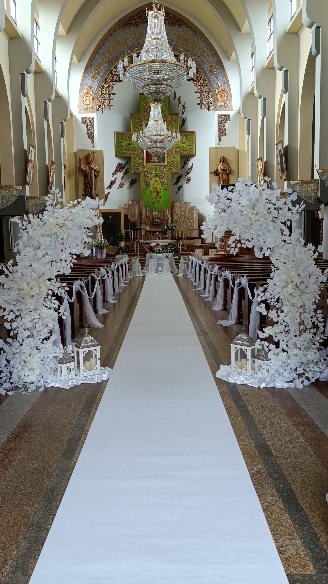 Uroczystość ślubna w kościele: biały dywan, dekoracje kwiatowe z białych gałązek, lampiony, wstążki na ławkach, oświetlenie z żyrandoli.