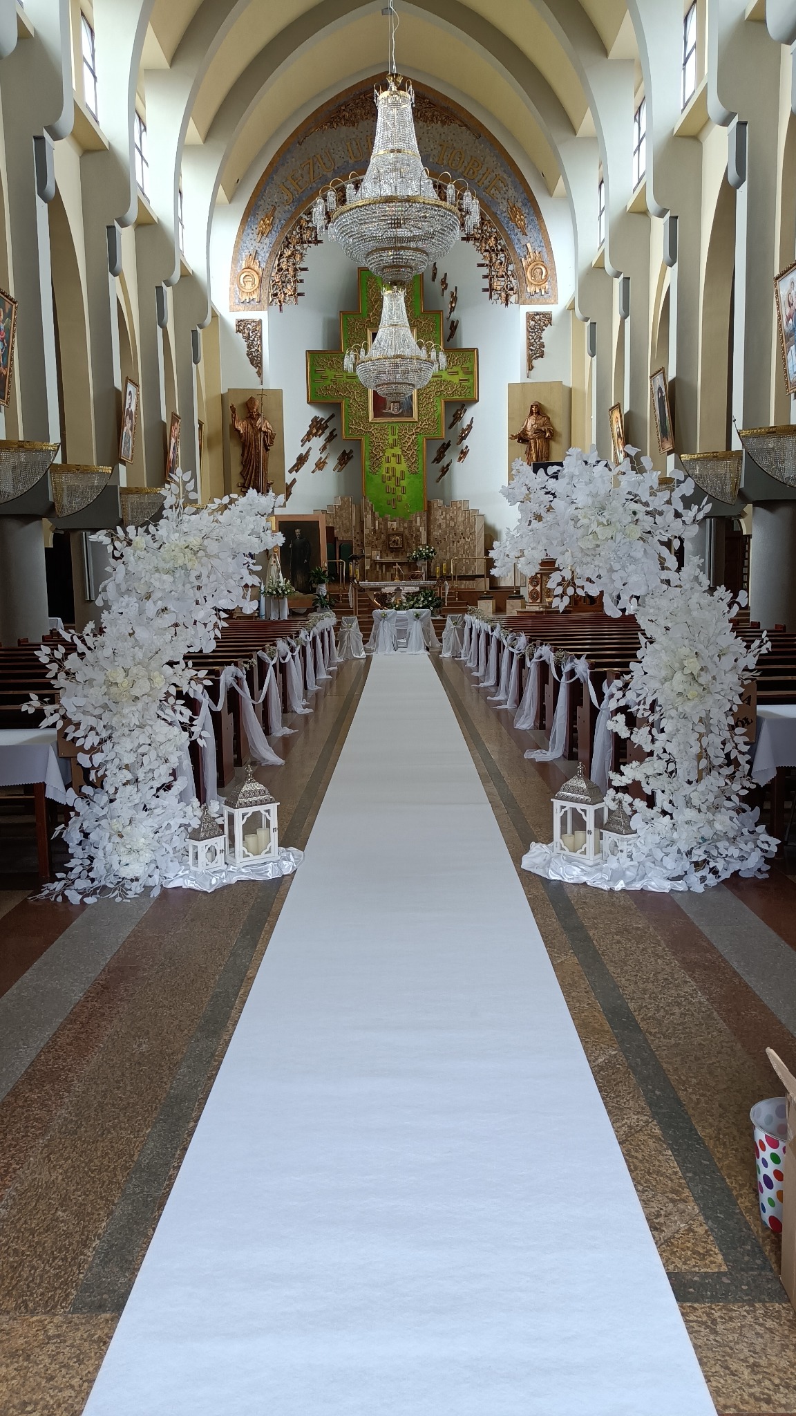 Uroczyste dekoracje w kościele: biały dywan, kwiatowe kompozycje przy ławkach, lampiony ze świecami, ozdobny żyrandol i zielony krzyż w tle.