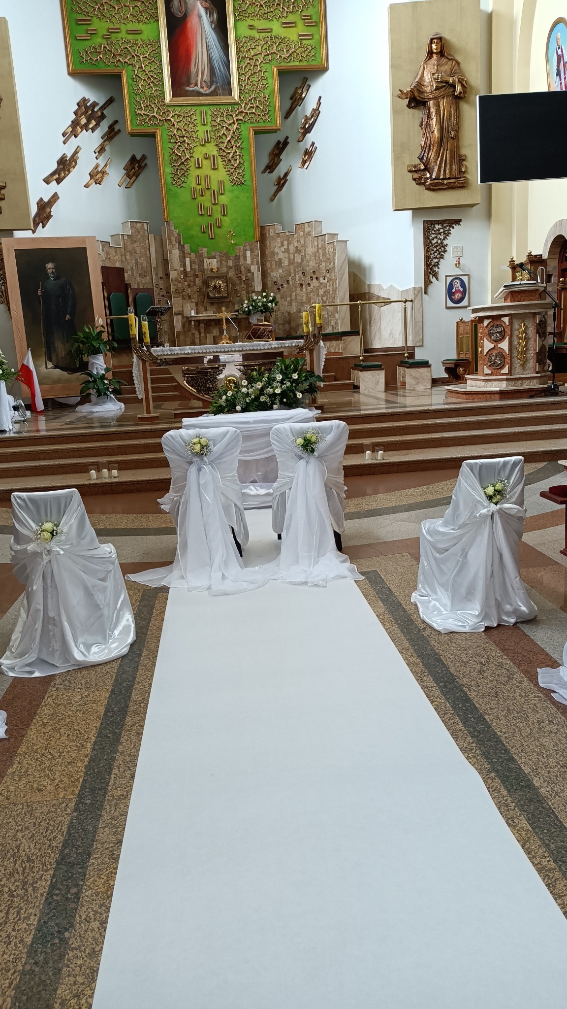 Uroczysta dekoracja kościoła przed ślubem: biały dywan, krzesła z białymi pokrowcami i kwiatowymi ozdobami, ołtarz w tle.