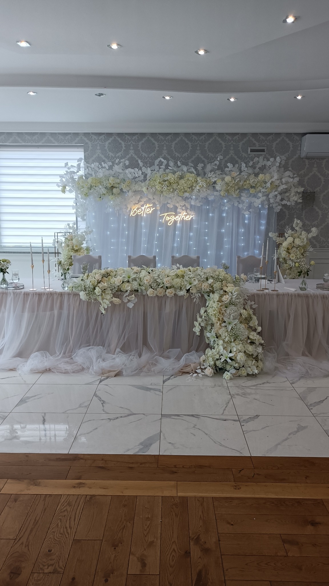 Uroczysta dekoracja stołu weselnego z białych kwiatów i neonowym napisem 'Better Together' na tle białej, zwiewnej tkaniny z delikatnym oświetleniem.