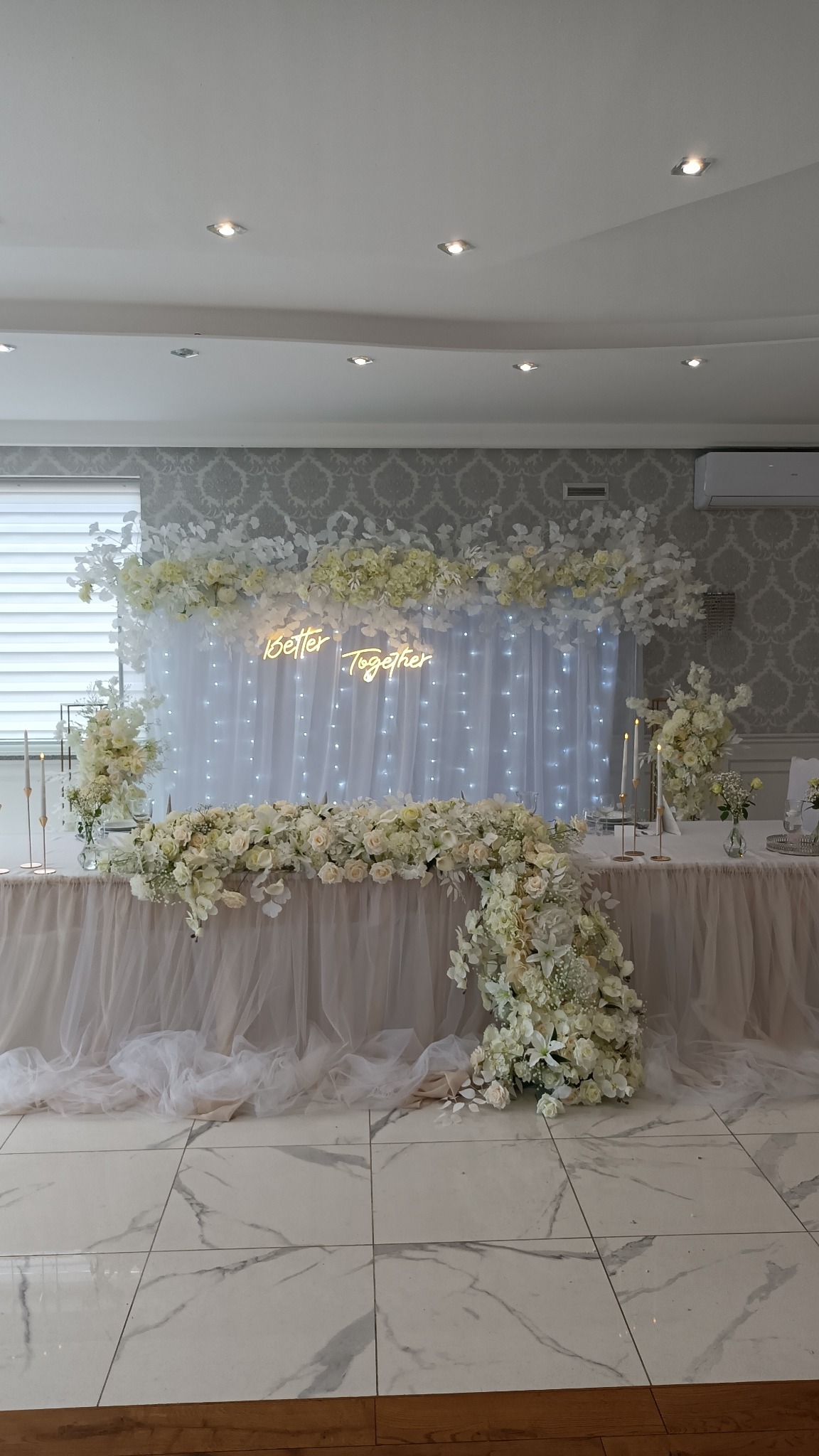 Uroczysty stół weselny udekorowany białymi kwiatami, podświetlony neonem z napisem 'Better Together', na tle białej zasłony z delikatnymi światełkami.