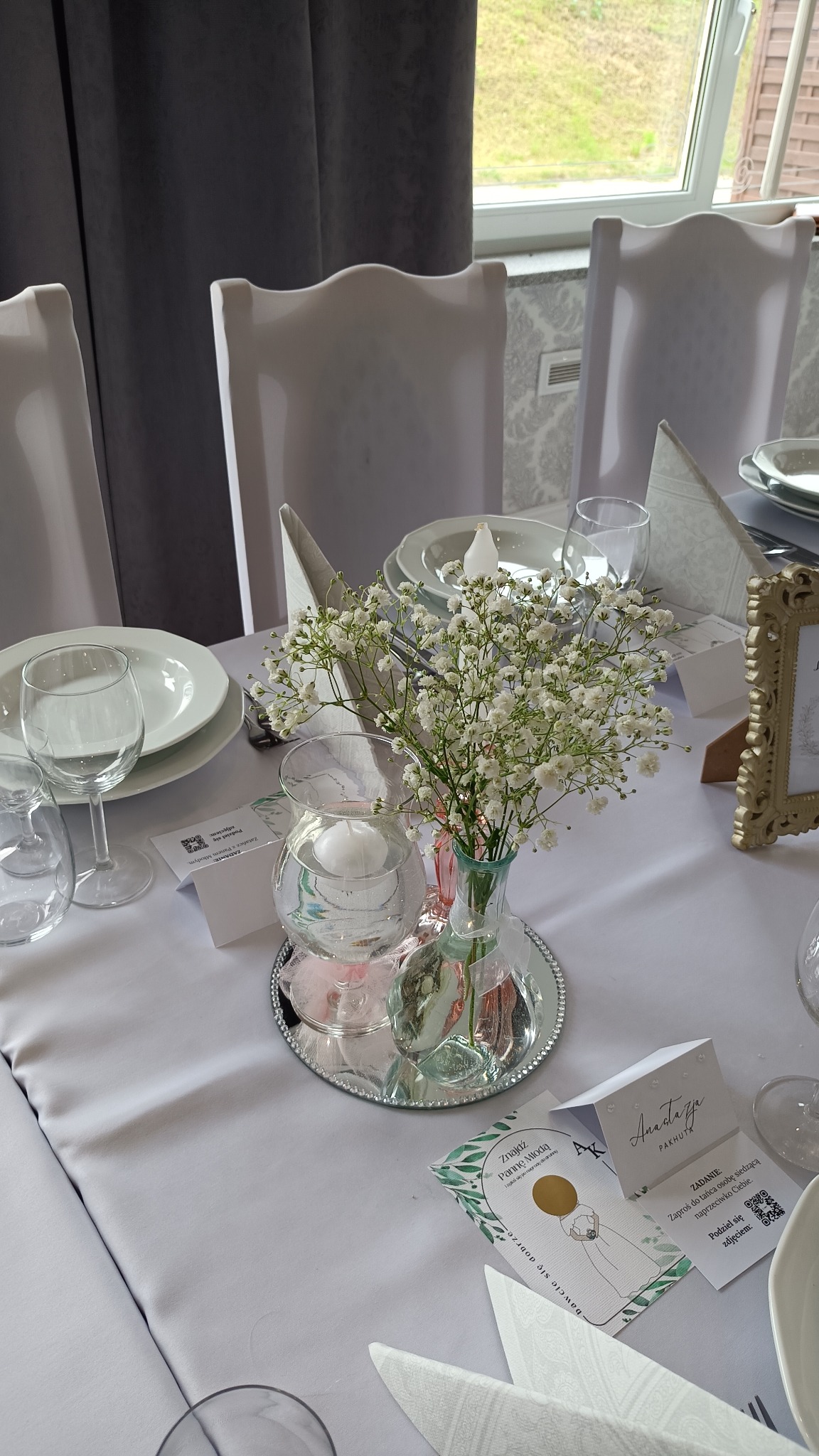 Dekoracja stołu weselnego z bukietem gipsówki w szklanym wazonie, świecami w szklanych kulach, białymi nakryciami i kartami z podziękowaniami dla gości.