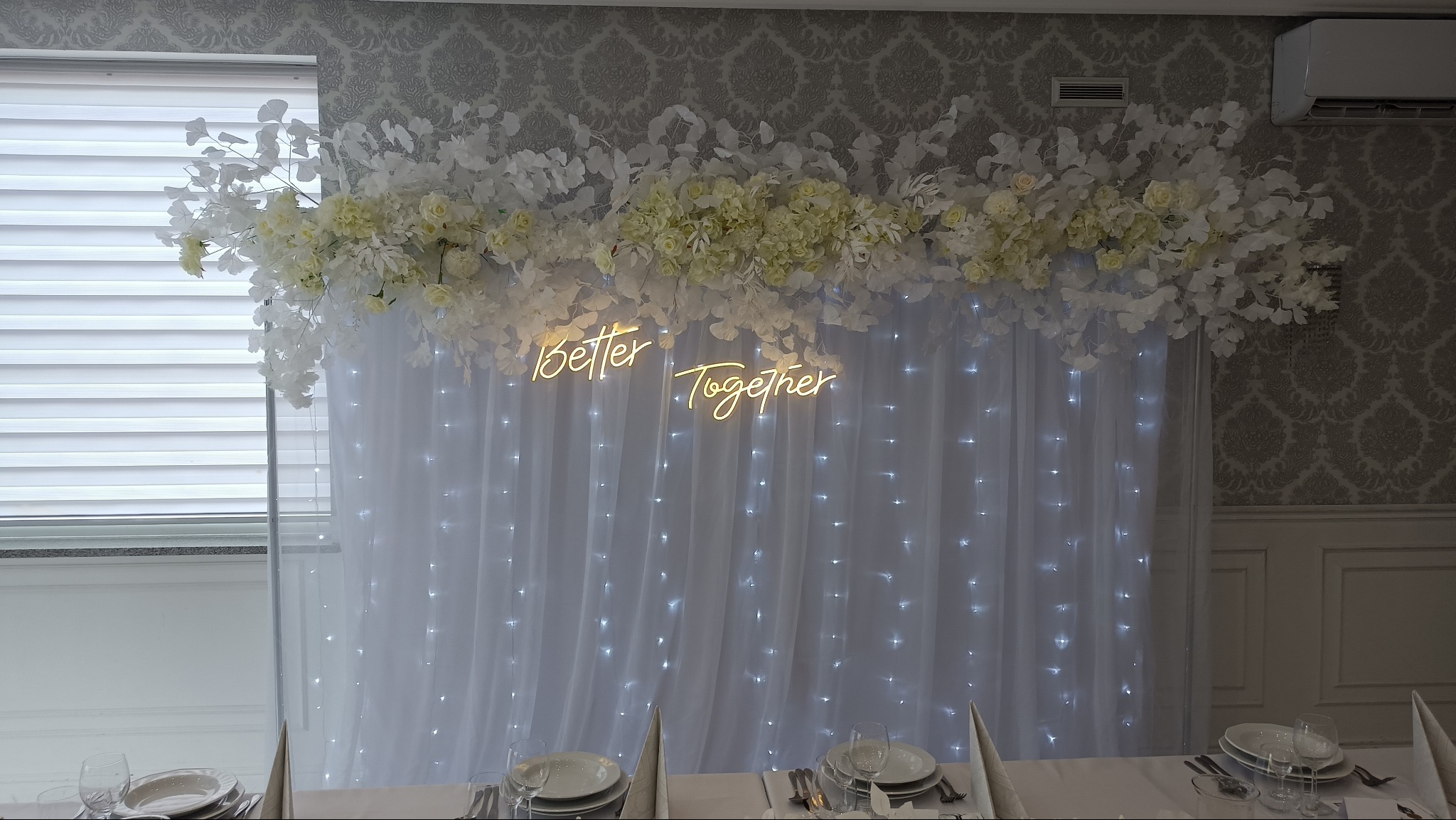 Dekoracja ślubna: białe kwiaty, liście i neonowy napis 'Better Together' na tle białych, podświetlanych lampek kurtynowych.