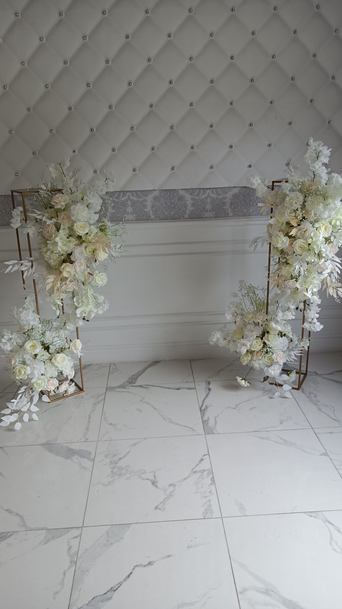 Dekoracja ślubna: złote, metalowe stelaże z kompozycjami z białych i kremowych kwiatów, na tle białej ściany z pikowaniem i marmurowej podłogi.