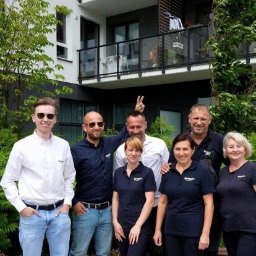 Greenic Sp z o.o. - Grupa siedmiu osób, prawdopodobnie ekipa sprzątająca, pozuje przed nowoczesnym budynkiem mieszkalnym z balkonami, ubrana w firmowe koszulki polo z logo.