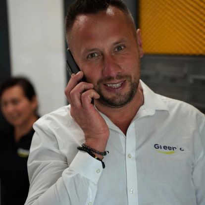 Uśmiechnięty mężczyzna w białej koszuli z logo firmy sprzątającej, rozmawia przez telefon. W tle widoczna inna osoba.