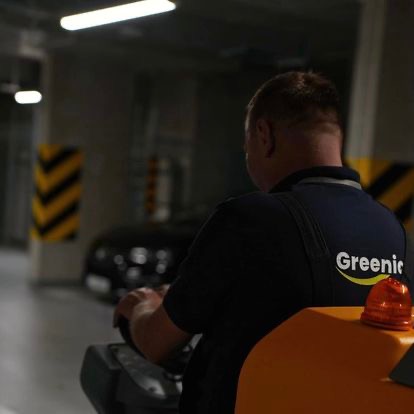 Pracownik firmy sprzątającej Greenic obsługuje szorowarkę w garażu podziemnym, widoczny fragment pojazdu i oznaczenia ostrzegawcze na filarze.