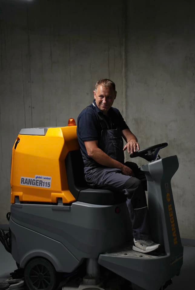 Uśmiechnięty mężczyzna w kombinezonie siedzi na żółto-szarej maszynie czyszczącej Ghibli Ranger 115 na tle betonowej ściany.