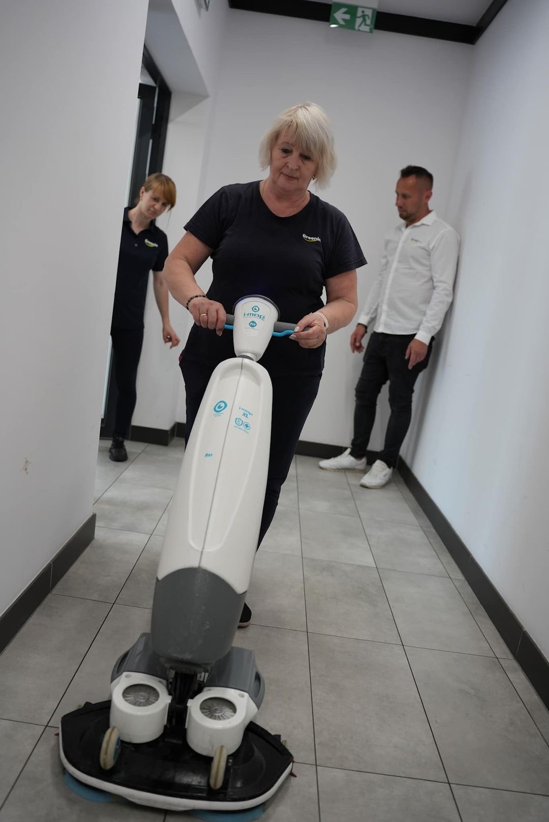 Kobieta obsługuje profesjonalną maszynę do czyszczenia podłóg w korytarzu biurowym, widoczni dwaj pracownicy w tle.