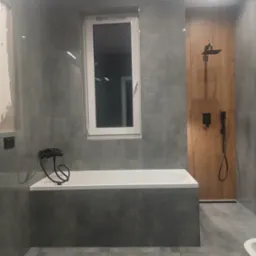 Wykończona łazienka z szarymi płytkami, białą wanną i czarną armaturą. Widoczna drewnopodobna ściana z deszczownicą. Fragment ściany przygotowany do położenia gładzi.