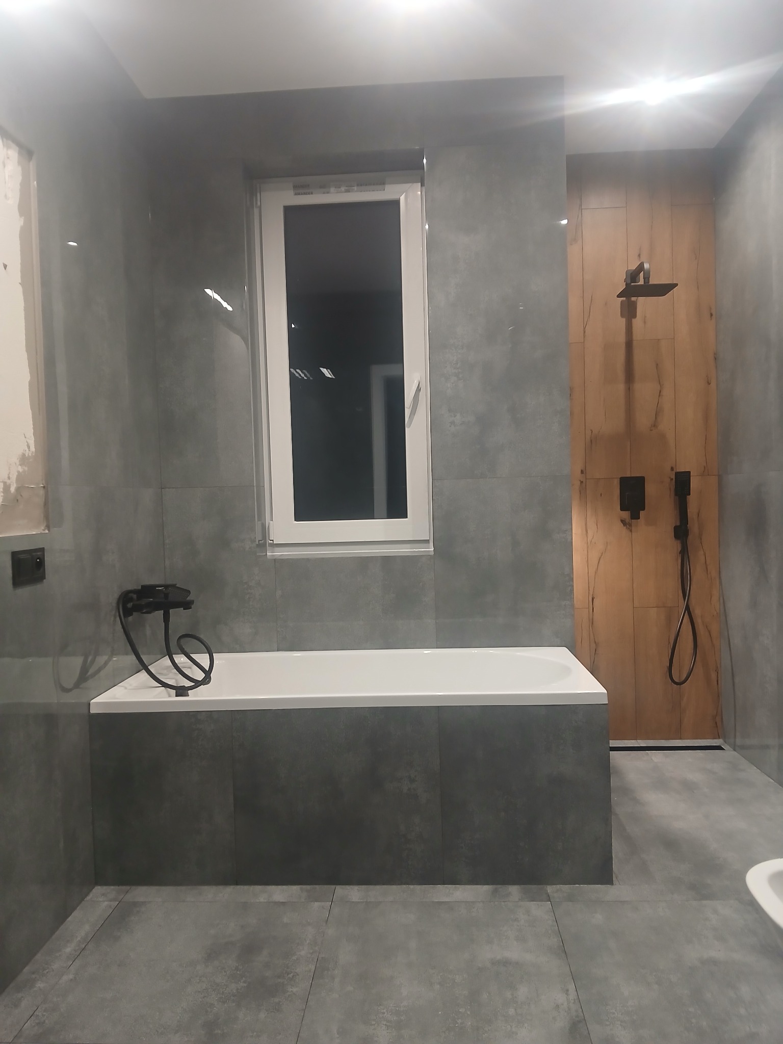 Wykończona łazienka z szarymi płytkami, białą wanną i czarną armaturą. Widoczna drewnopodobna ściana z deszczownicą. Fragment ściany przygotowany do położenia gładzi.