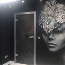 Ciemne wnętrze łazienki z kabiną prysznicową z przyciemnianego szkła i dekoracyjnym malowidłem na ścianie przedstawiającym portret kobiety w koronie z kryształów.