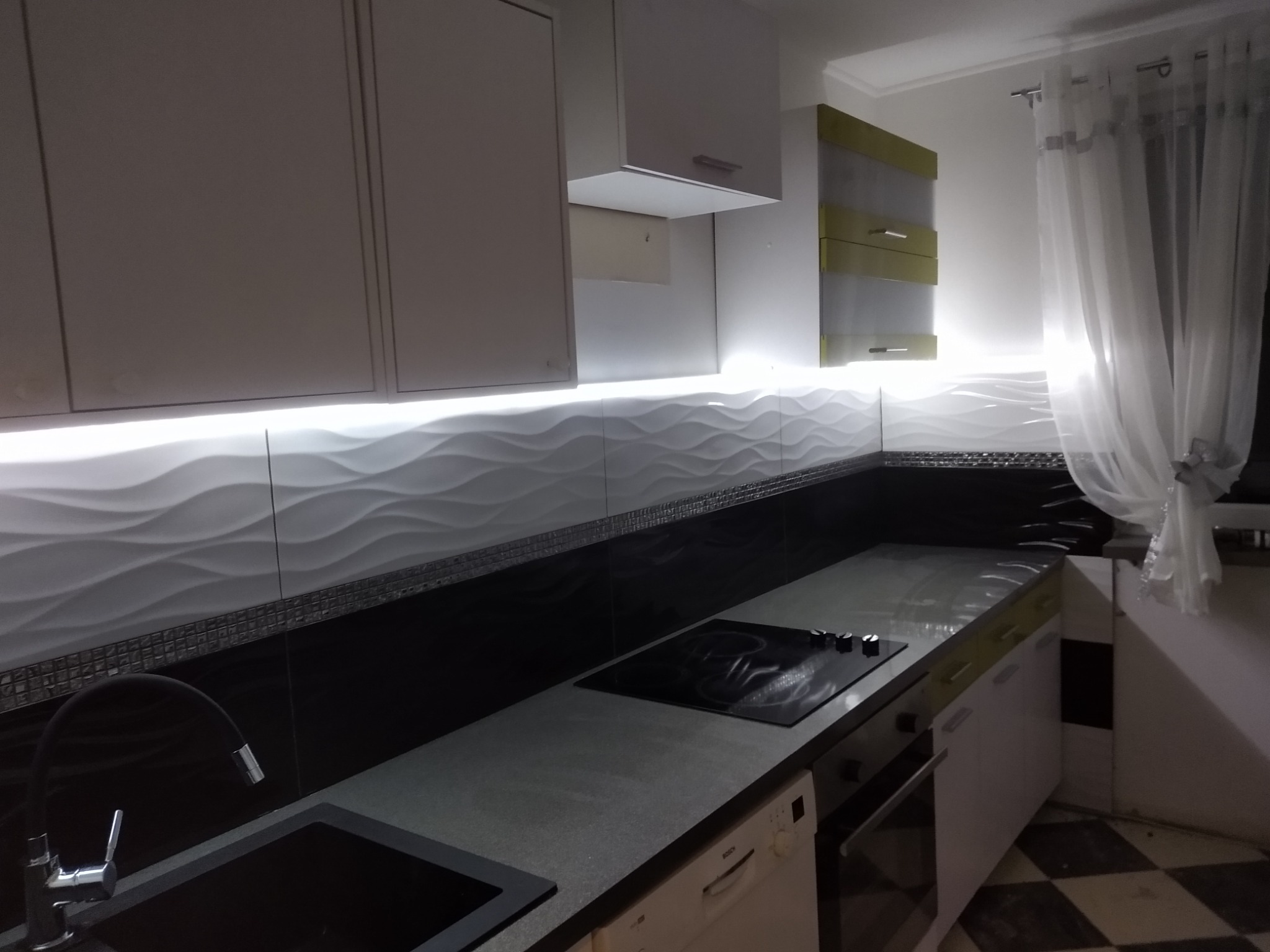 Nowoczesna kuchnia z białymi szafkami, podświetlanymi panelami ściennymi o falistej strukturze, czarnym blatem i płytą grzewczą oraz podłogą w szachownicę.