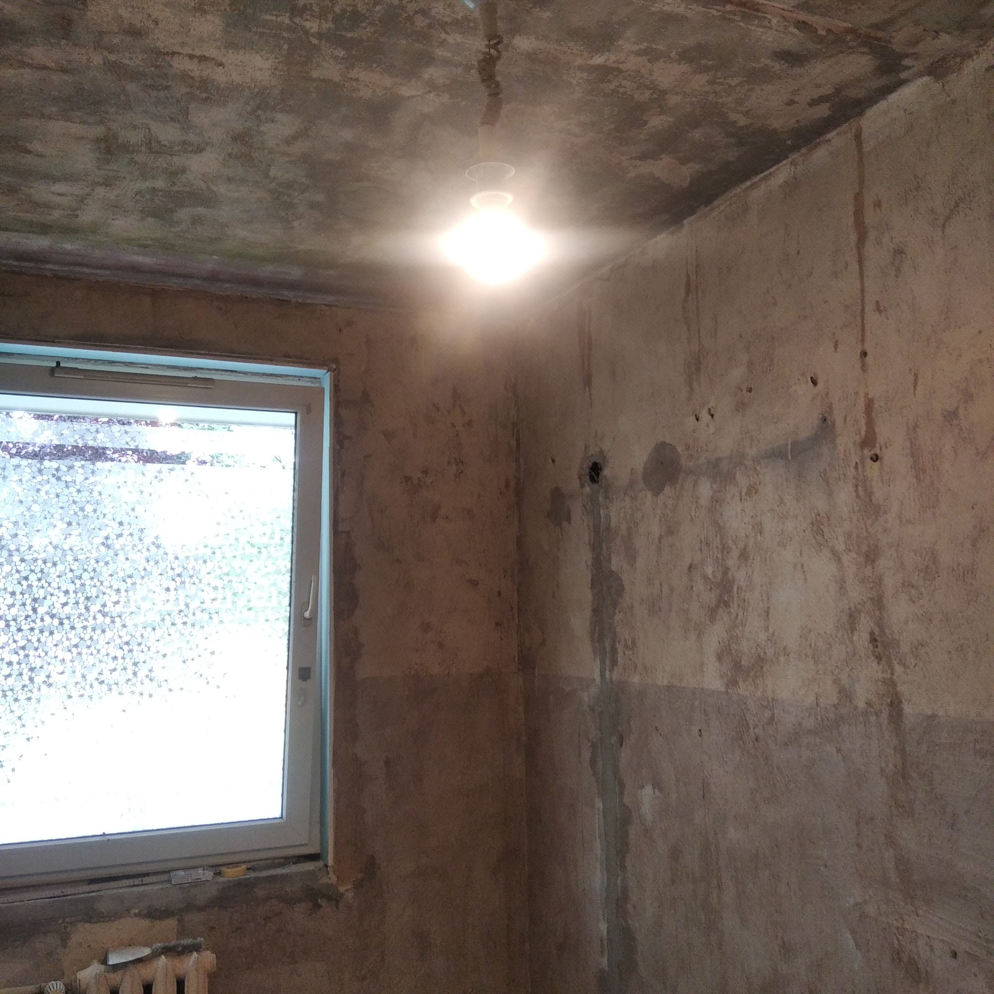 Surowe ściany i sufit w pokoju przed remontem, widoczne ślady po wcześniejszych instalacjach, pojedyncza żarówka oświetla wnętrze, okno z ozdobną folią. Grzejnik częściowo widoczny pod oknem.