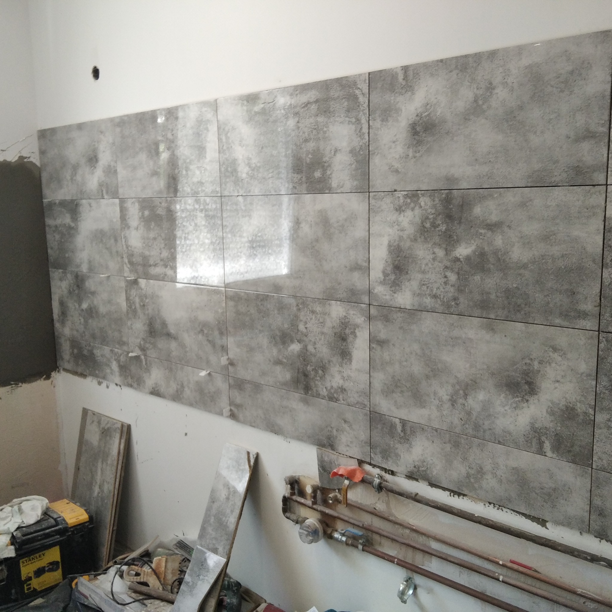 Ściana w trakcie układania szarych, matowych płytek w kuchni, widoczne rury instalacji wodnej, skrzynka narzędziowa Stanley i resztki materiałów budowlanych.