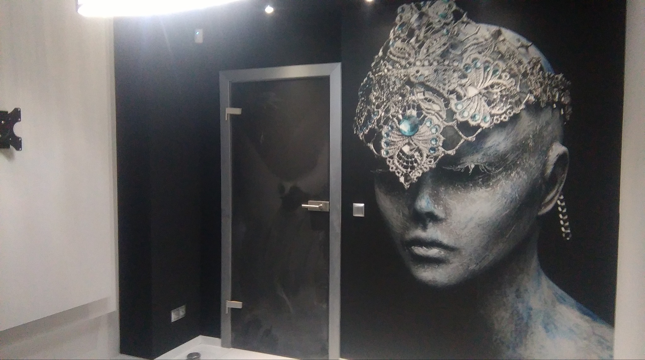 Ciemne wnętrze łazienki z kabiną prysznicową z przyciemnianego szkła i dekoracyjnym malowidłem na ścianie przedstawiającym portret kobiety w koronie z kryształów.