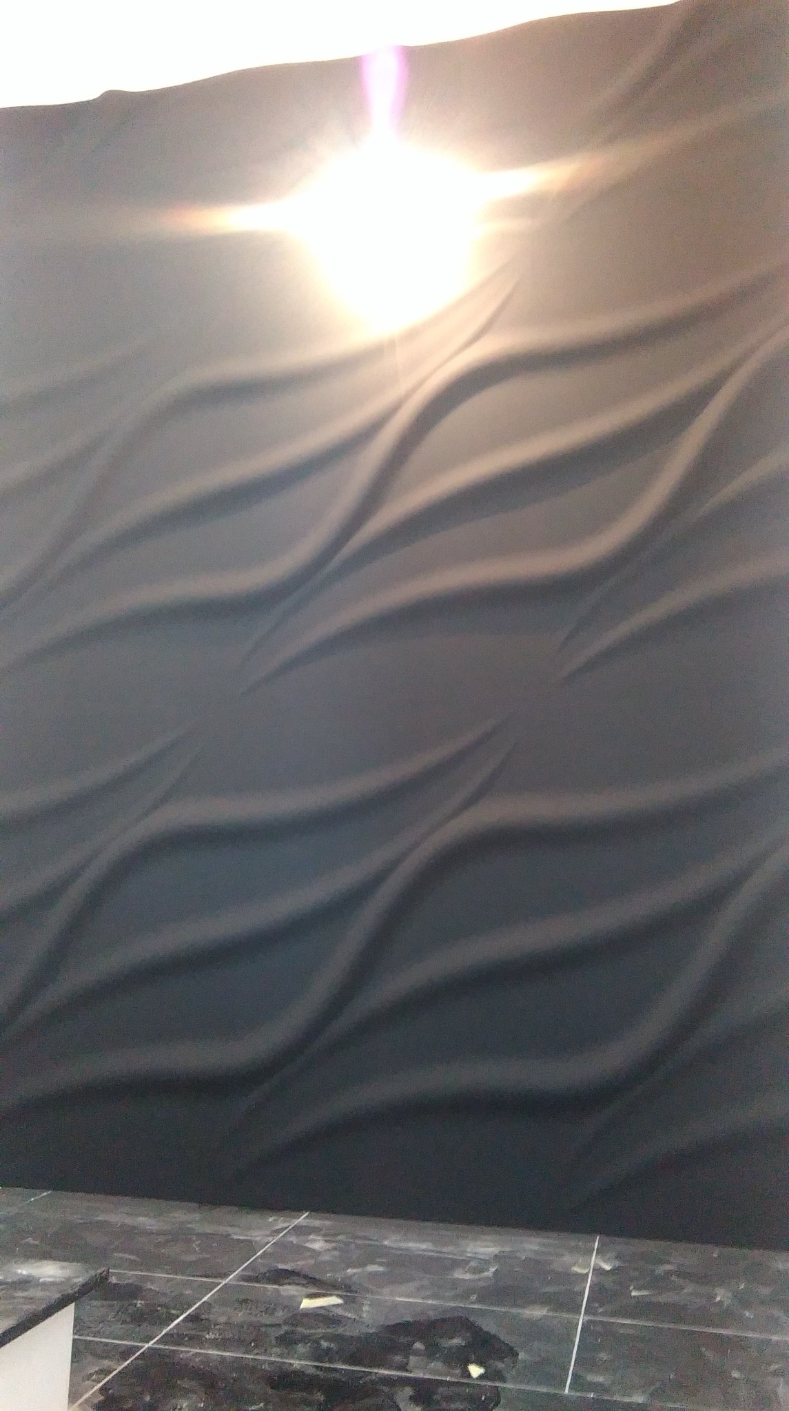 Ciemnoszara ściana z trójwymiarowym, falistym wzorem, oświetlona od góry, z widoczną podłogą wyłożoną ciemnymi płytkami.