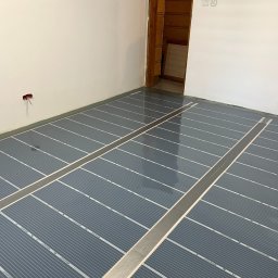 Eneka sc - Szare maty grzewcze rozłożone na podłodze w pomieszczeniu z białymi ścianami i drewnianymi drzwiami, gotowe do montażu