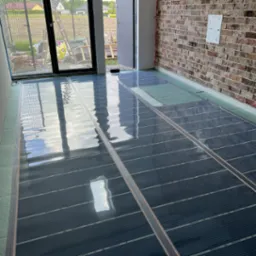 Instalacja mat grzewczych w nowym domu, widoczne panele grzewcze na podłodze, przygotowane do zalania wylewką, ściana z cegły w tle.