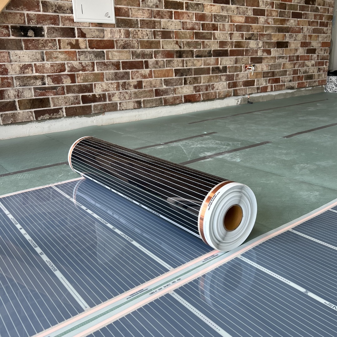 Rolka rozwijanej elektrycznej folii grzewczej marki BRAVE na podłodze, widoczna ceglana ściana w tle.