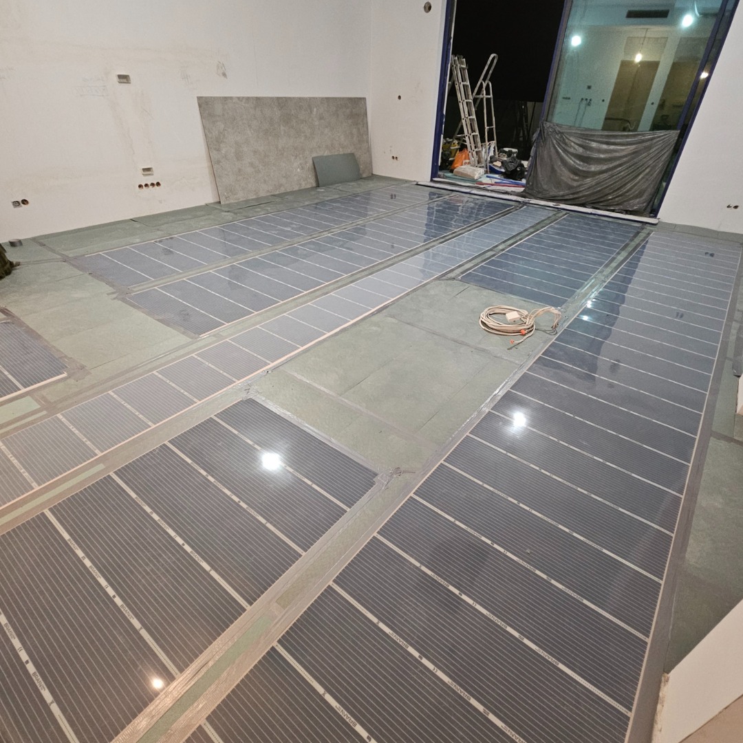 Rozłożone sekcje systemu ogrzewania podłogowego, prawdopodobnie elektryczne maty grzewcze, przed wylaniem wylewki samopoziomującej w nowym pomieszczeniu z dużym oknem balkonowym.