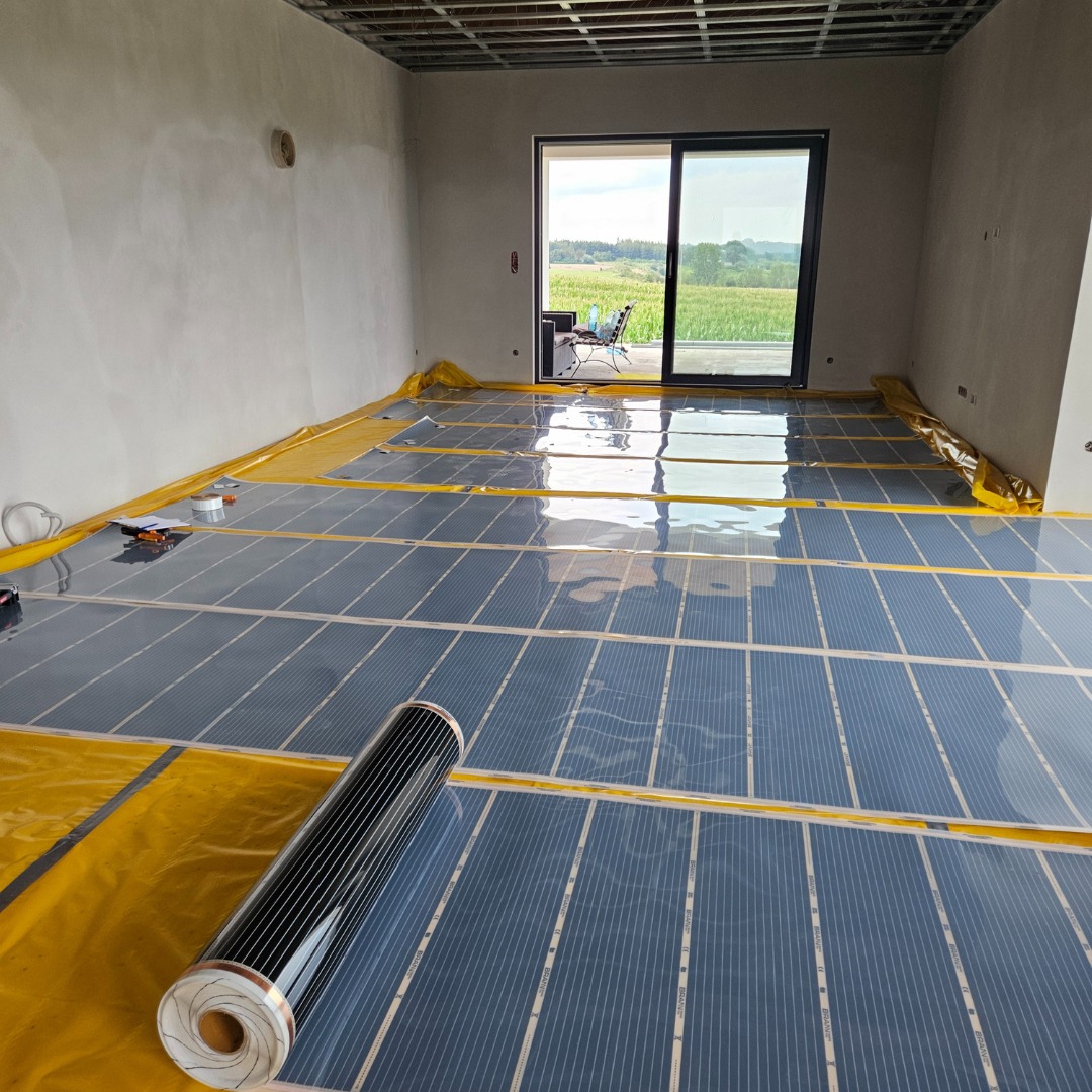 Rozłożona na żółtej folii podłogowa mata grzewcza z widocznym zrolowanym fragmentem, przygotowana do montażu w surowym pomieszczeniu z widokiem na zielone pola za oknem.