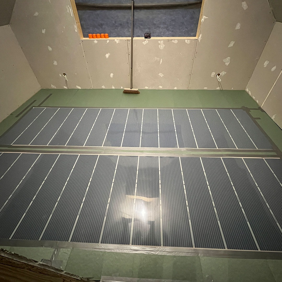 Montaż elektrycznego ogrzewania podłogowego w pomieszczeniu ze skośnym sufitem i oknem dachowym, widoczne maty grzewcze rozłożone na zielonej płycie podłogowej, przygotowane do dalszych prac...