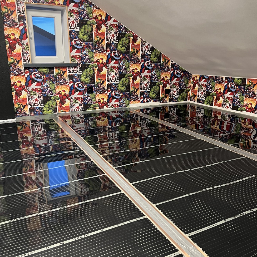 Instalacja czarnej maty grzewczej na poddaszu z tapetą w motywy komiksowe Marvela, widoczne srebrne listwy mocujące.