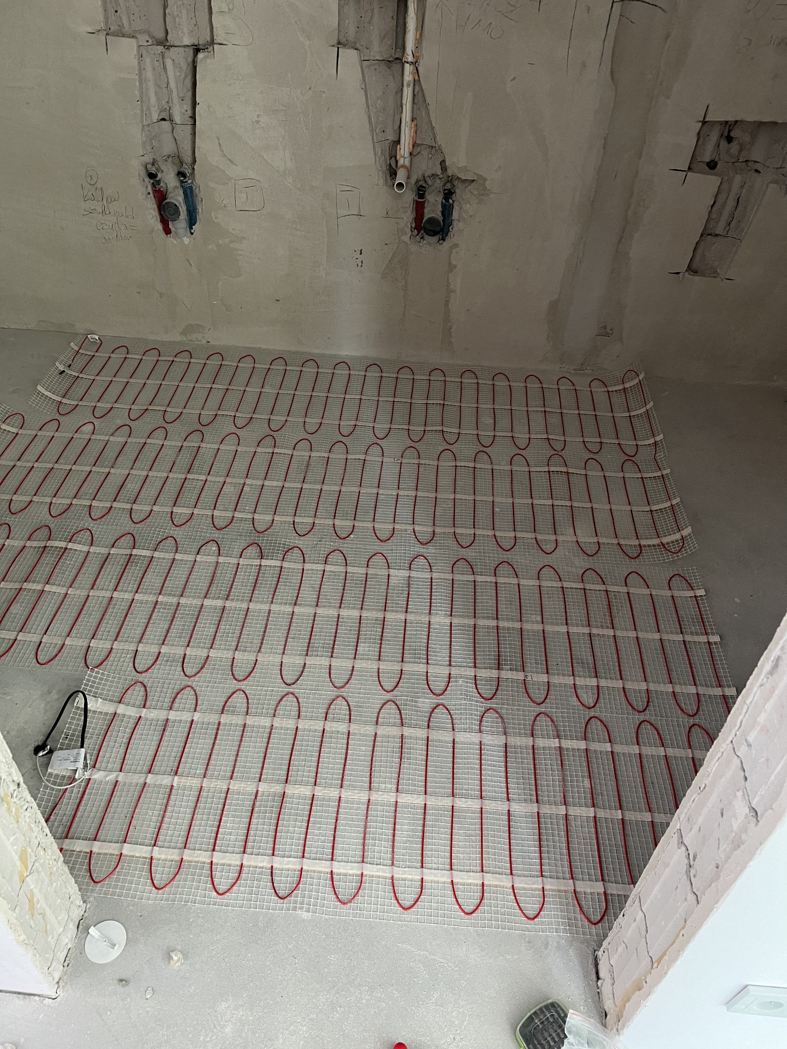 Rozłożona mata grzewcza z czerwonymi przewodami na białej siatce, widoczne rury instalacji na ścianie w surowym stanie.