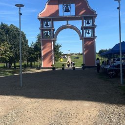 Usługi Transportowe Józef Basarab - Monumentalna brama z dzwonami i krzyżem na szczycie, widziana z perspektywy żwirowej drogi, prowadząca do zielonego wzgórza z krzyżem w tle, w słoneczny dzień.