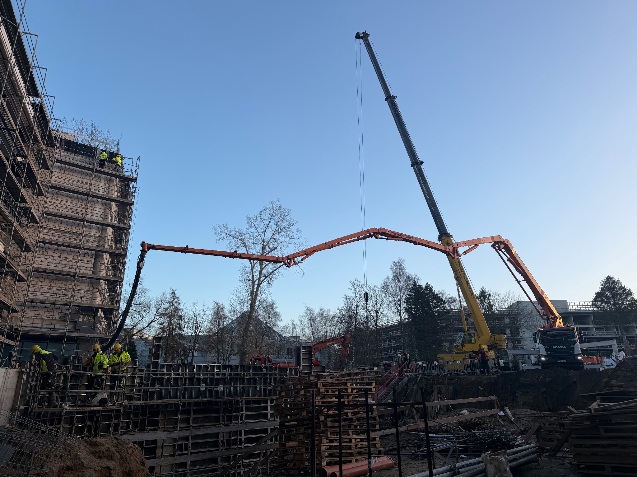Budowa w toku: pracownicy w żółtych kamizelkach, szalunki fundamentów, dźwig z pompą do betonu i koparka na tle drzew i częściowo ukończonego budynku.
