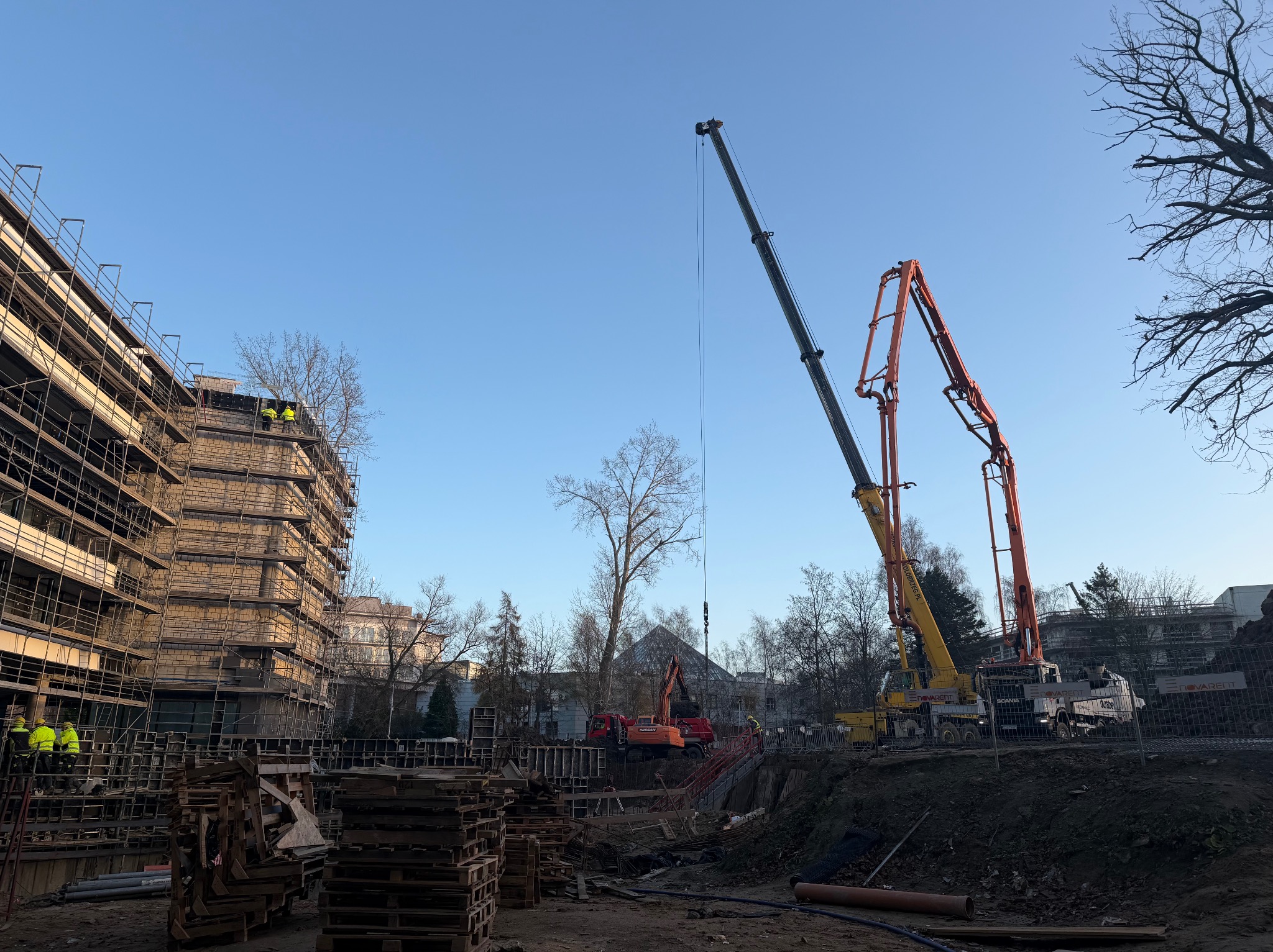 Budowa budynku w toku: widoczne rusztowania, dźwig, pompy do betonu, wykop i palety. Na rusztowaniach i w wykopie pracownicy w żółtych kamizelkach. Błękitne niebo w tle.