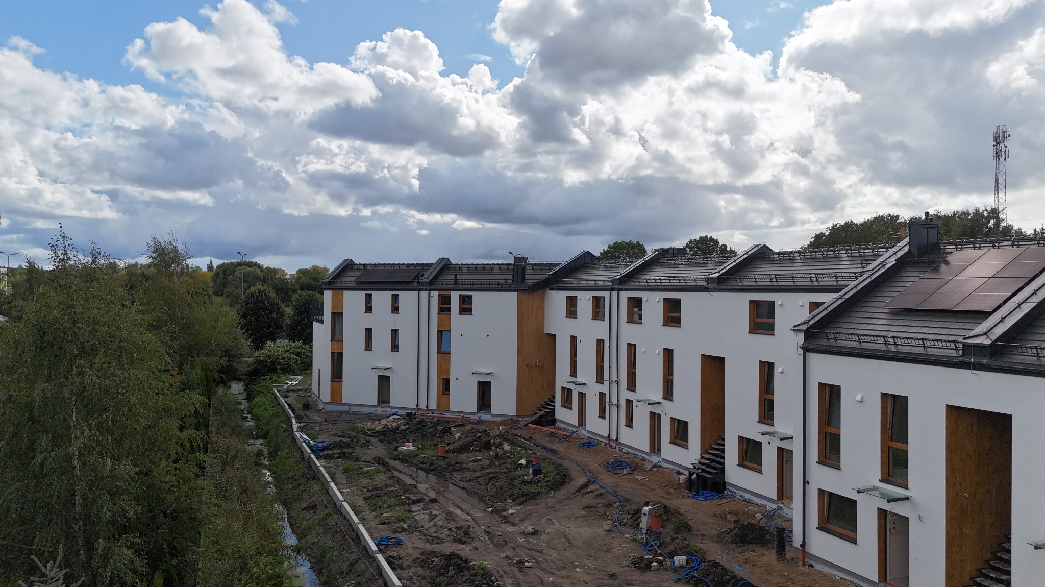 Nowoczesne, białe domy szeregowe z panelami słonecznymi na dachu, widoczne z góry, z przygotowanym terenem pod ogródki i instalacjami na zewnątrz.