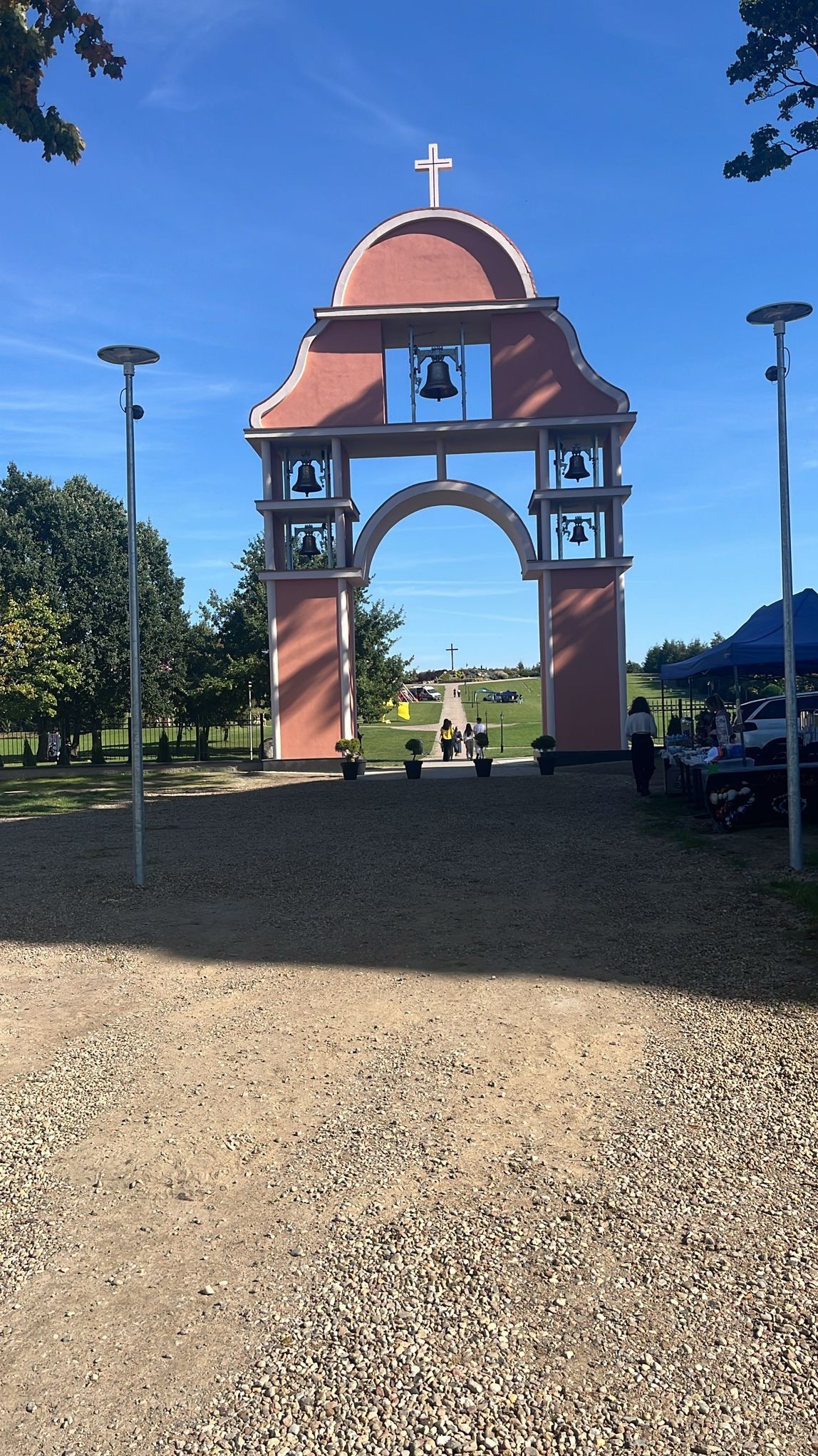 Monumentalna brama z dzwonami i krzyżem na szczycie, widziana z perspektywy żwirowej drogi, prowadząca do zielonego wzgórza z krzyżem w tle, w słoneczny dzień.
