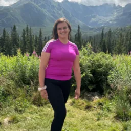 Uśmiechnięta kobieta w różowej koszulce na tle górskiego krajobrazu z lasem i łąką. Słoneczny dzień, widoczne chmury.