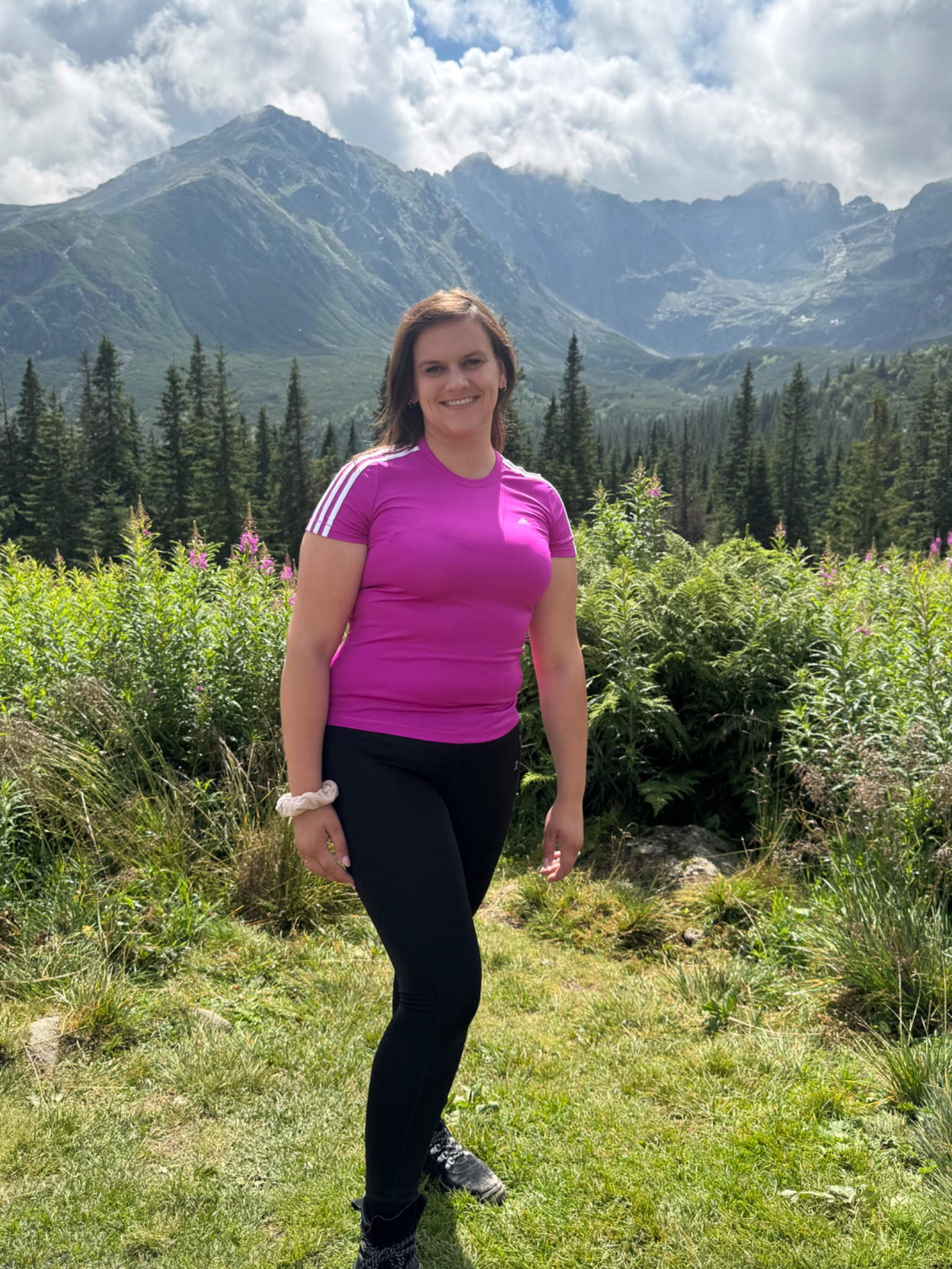 Uśmiechnięta kobieta w różowej koszulce na tle górskiego krajobrazu z lasem i łąką. Słoneczny dzień, widoczne chmury.