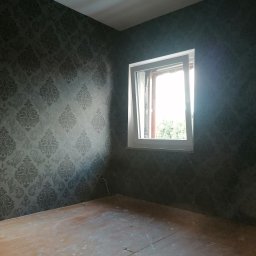D&P-BUD Łapiński Patryk - Pokój po malowaniu ścian na ciemny kolor z wzorzystą tapetą, widoczne drewniane podłogi w trakcie renowacji oraz okno z widokiem na zieleń.