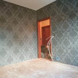 D&P-BUD Łapiński Patryk - Pokój w trakcie remontu z tapetą w szaro-srebrne ornamenty, widoczne wystające kable elektryczne i fragment sąsiedniego pomieszczenia z materiałami budowlanymi.