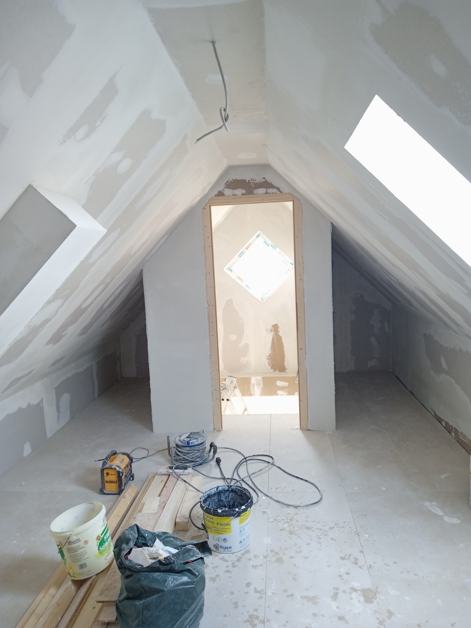 Poddasze w trakcie wykańczania: ściany i skosy pokryte szpachlą, widoczne narzędzia, kable, wiaderka z farbą i resztki materiałów budowlanych na podłodze.