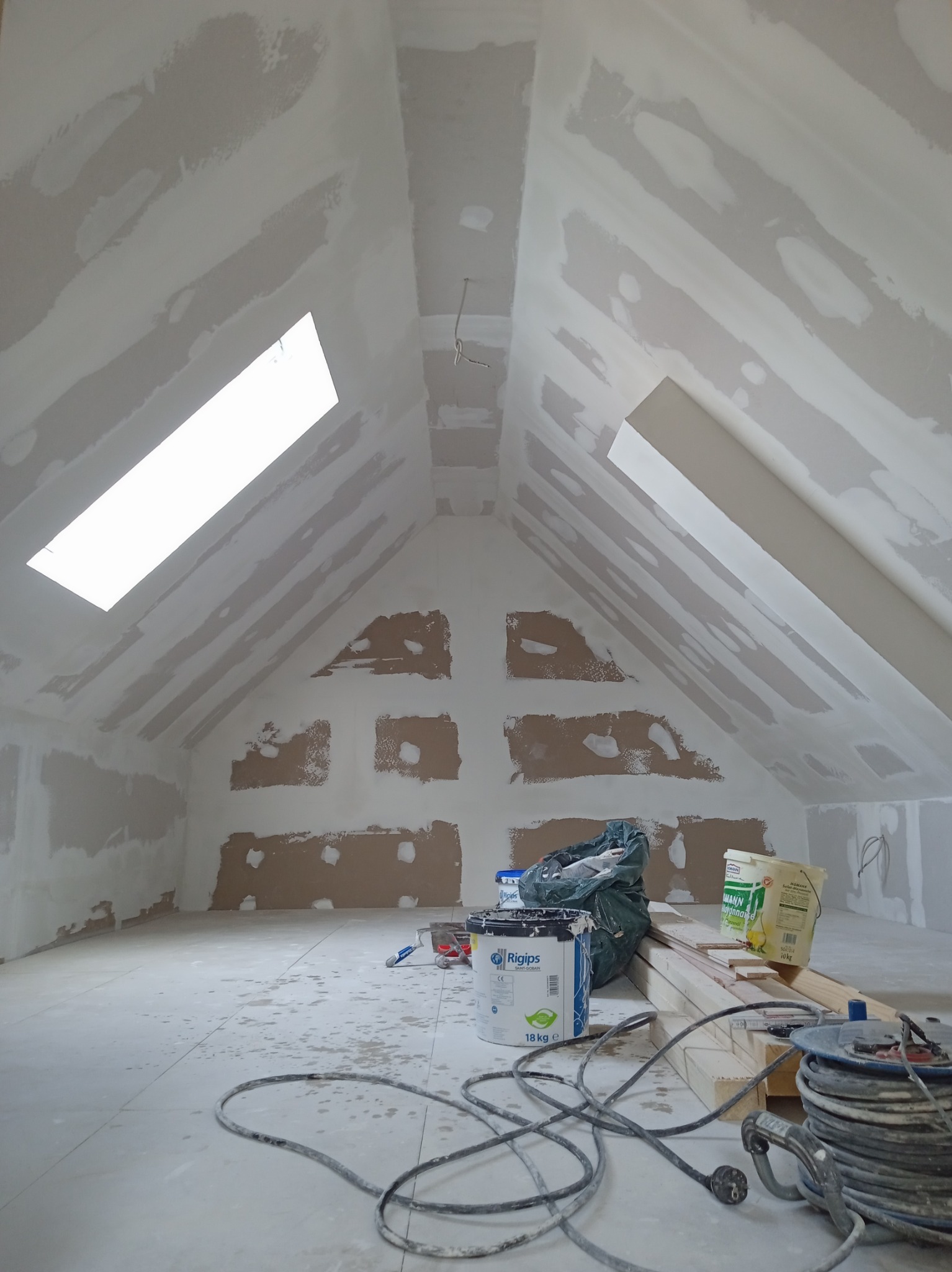 Poddasze w trakcie wykańczania: widoczne płyty karton-gips, częściowo zaszpachlowane, okno dachowe, puszka z gipsem Rigips, kabel i wiadro z klejem na podłodze.