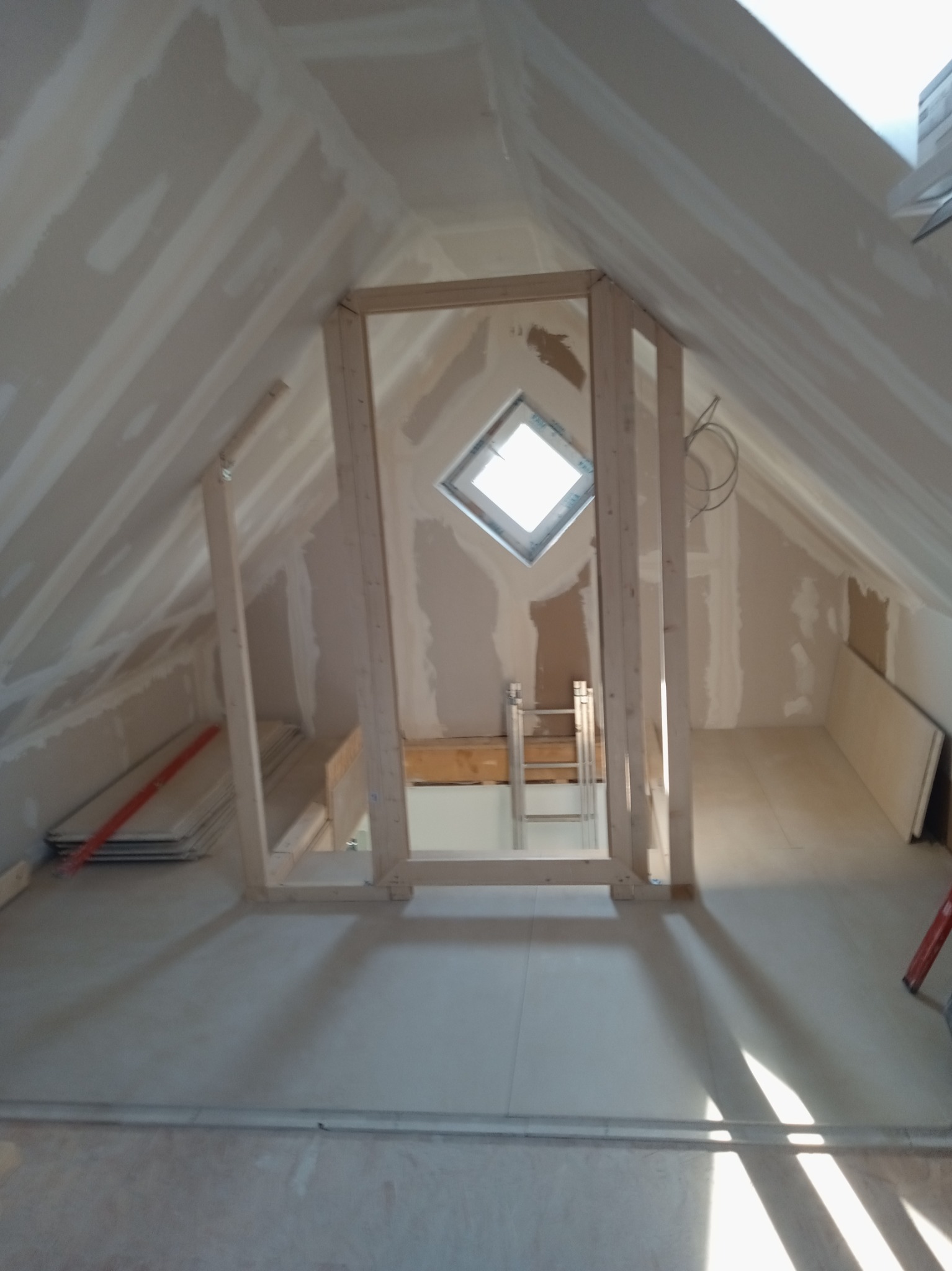 Poddasze w trakcie adaptacji: surowe ściany z płyt kartonowo-gipsowych, drewniana konstrukcja pod zabudowę, okno dachowe w kształcie rombu, widoczne przewody elektryczne.