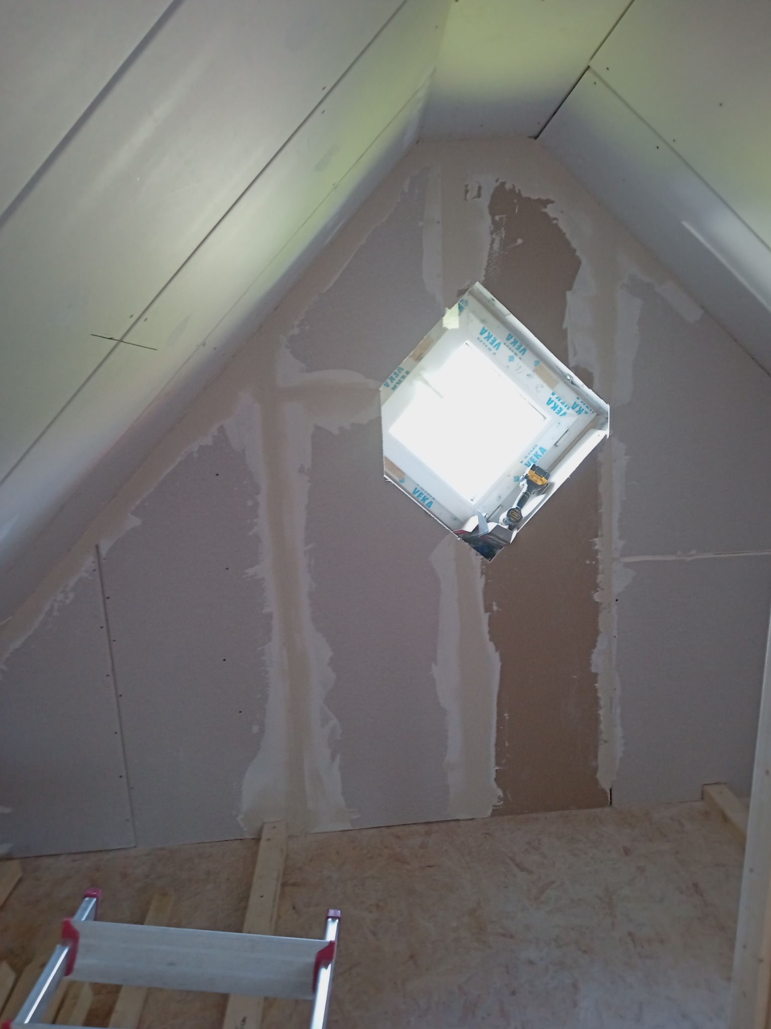 Montaż płyty karton gips w skośnym poddaszu z oknem dachowym, widoczne szpachlowane łączenia, drabina i narzędzia.