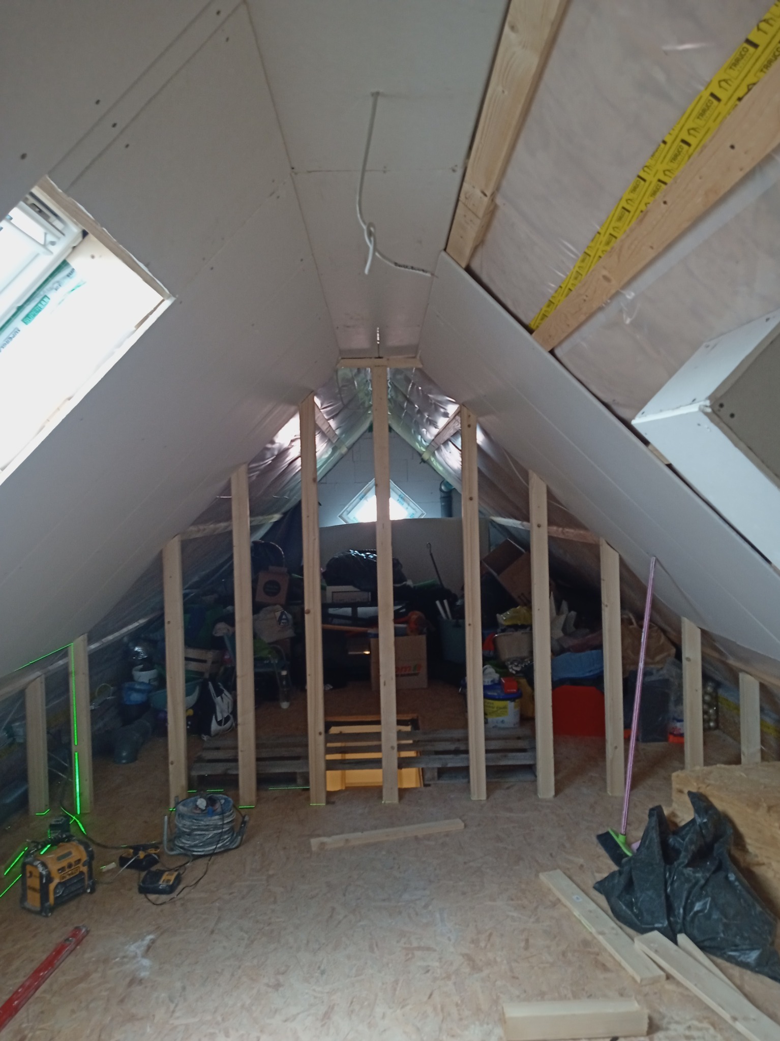 Poddasze w trakcie adaptacji: drewniana konstrukcja szkieletowa, częściowo zamontowane płyty gipsowo-kartonowe, widoczne narzędzia budowlane i laser krzyżowy wyznaczający poziom, w tle zgromadzone...