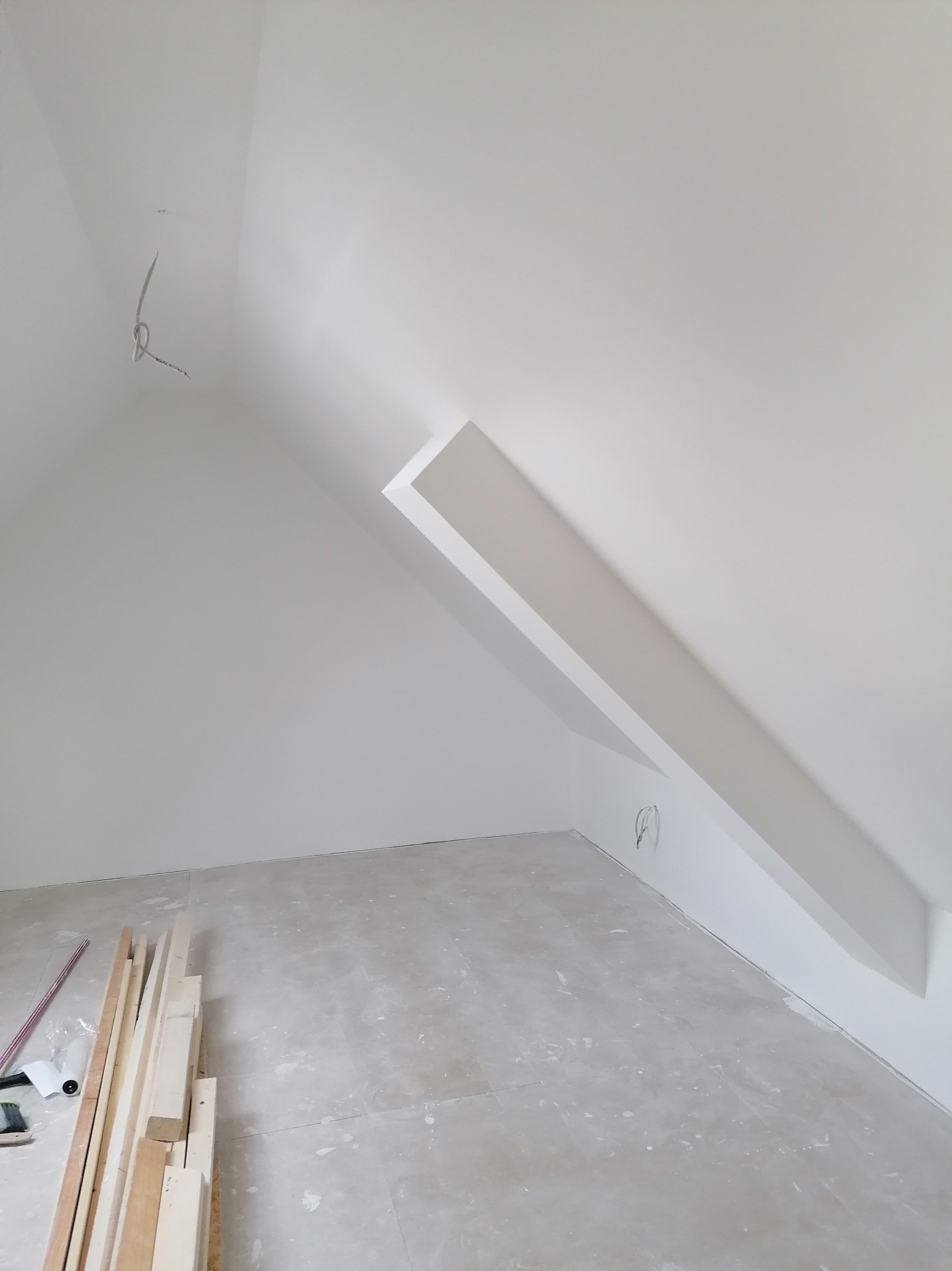 Poddasze w trakcie prac wykończeniowych, widoczne drewniane listwy przypodłogowe, przewody elektryczne wystające ze ściany oraz świeżo pomalowane ściany na biało.