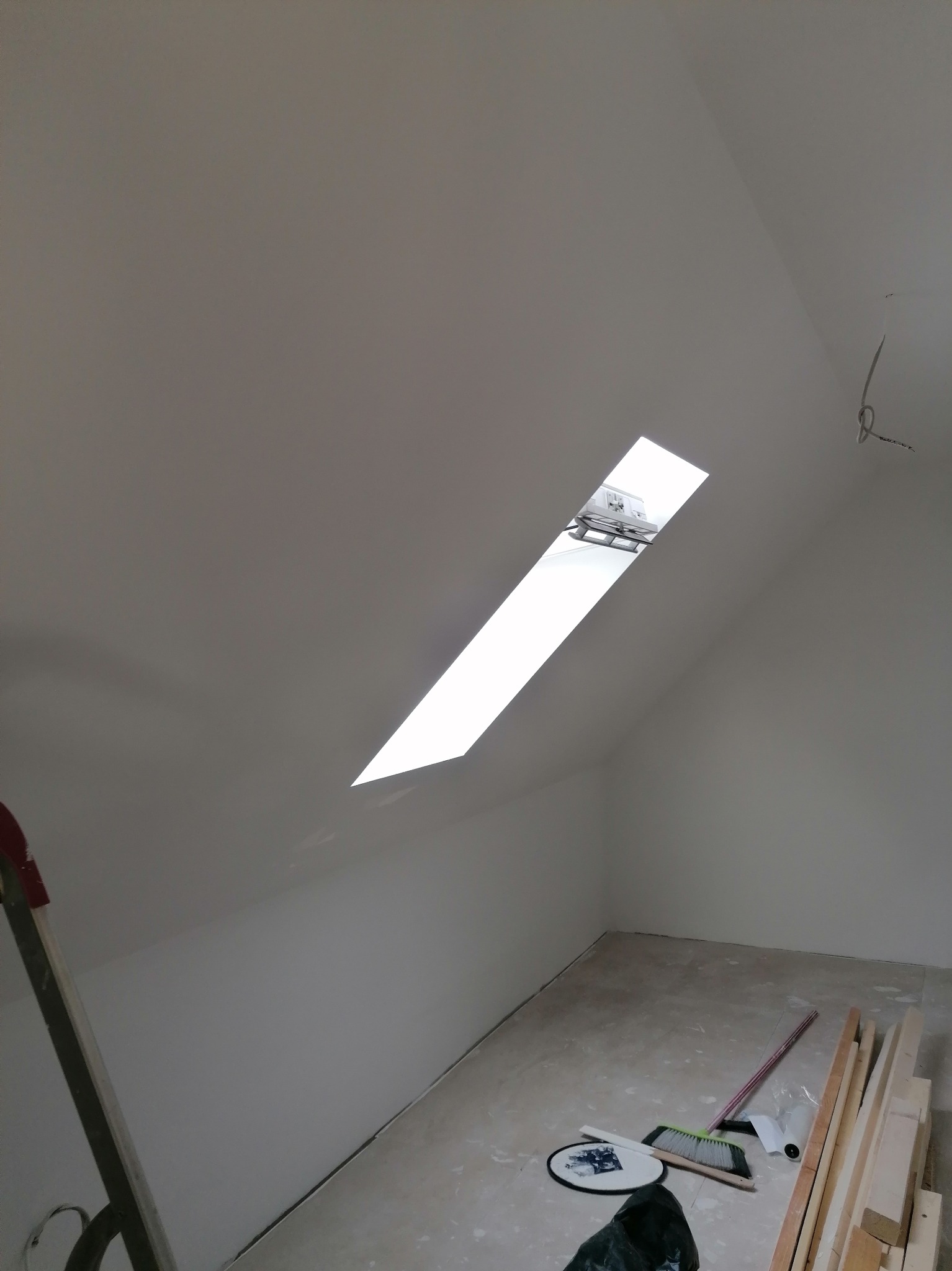 Poddasze po malowaniu na biało, widoczne okno dachowe, drabina oparta o ścianę, na podłodze szczotka, listwy przypodłogowe i inne narzędzia.