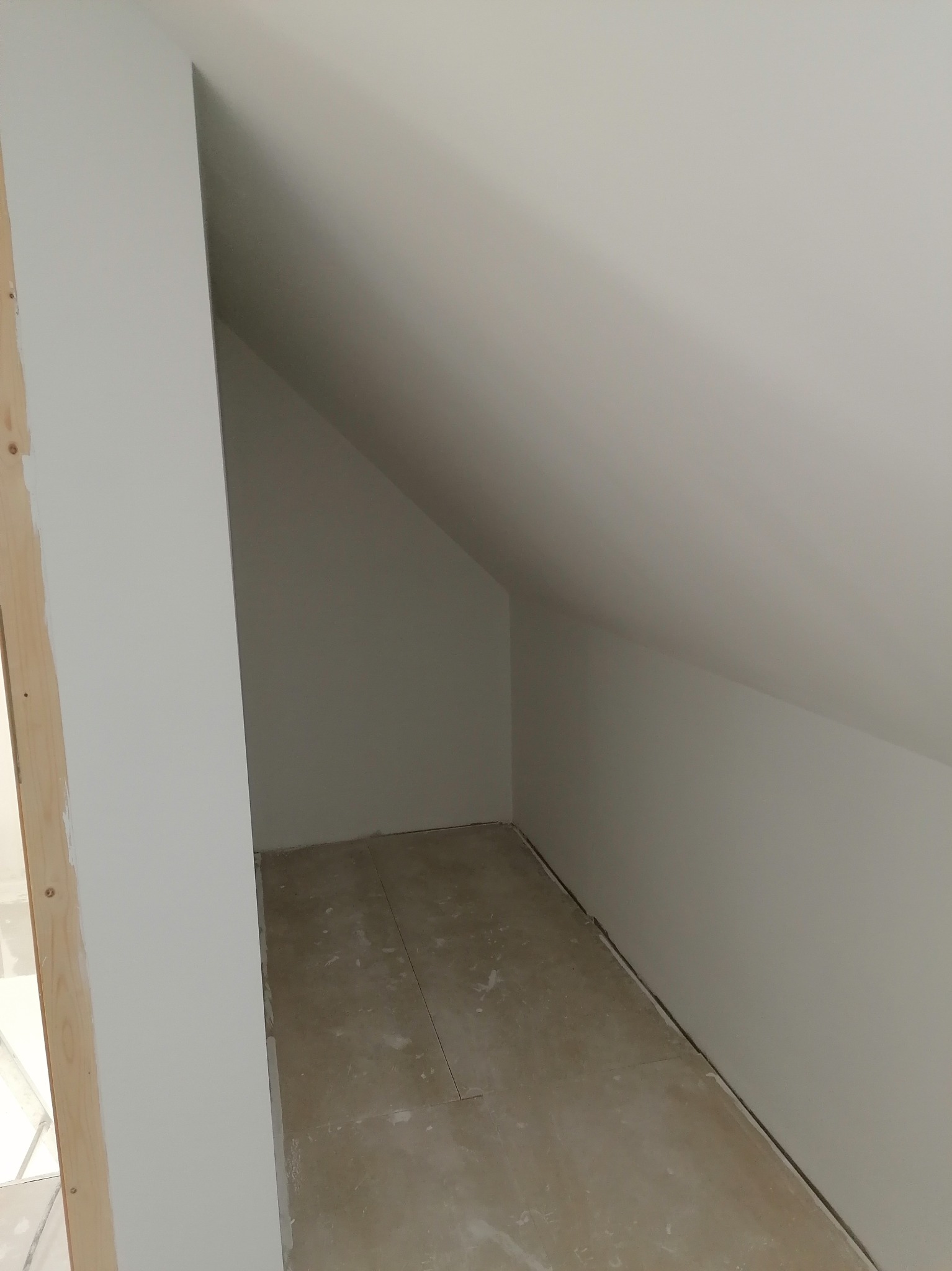 Pomalowane na biało wnętrze poddasza ze skośnym sufitem, widoczne ślady po remoncie na podłodze, drewniana rama w lewym rogu.