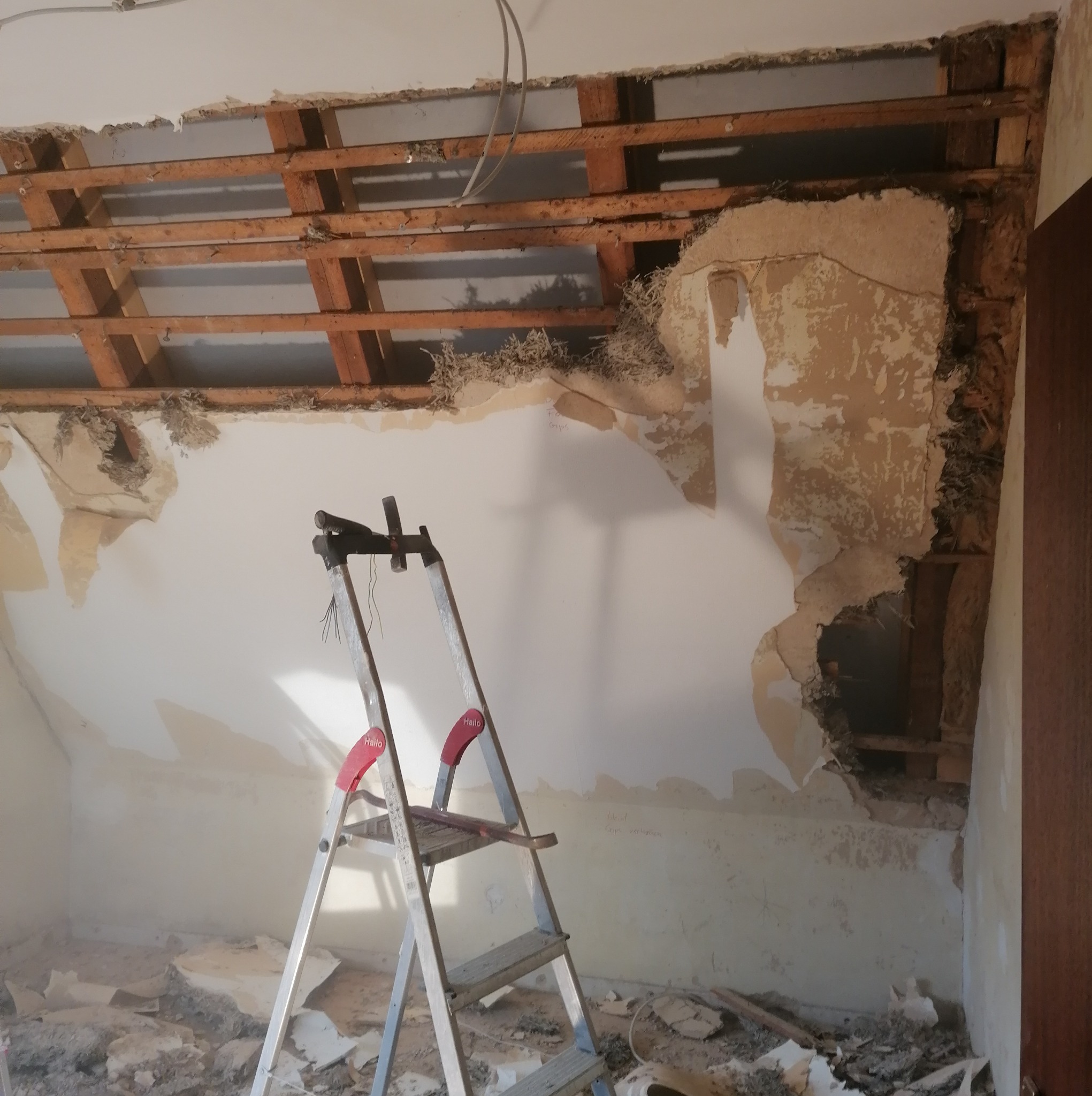 Poddasze w trakcie remontu, widoczne odsłonięte drewniane belki konstrukcyjne, fragmenty starej izolacji, ściany z resztkami tapet i tynku, drabina aluminiowa z narzędziami, gruz na podłodze.