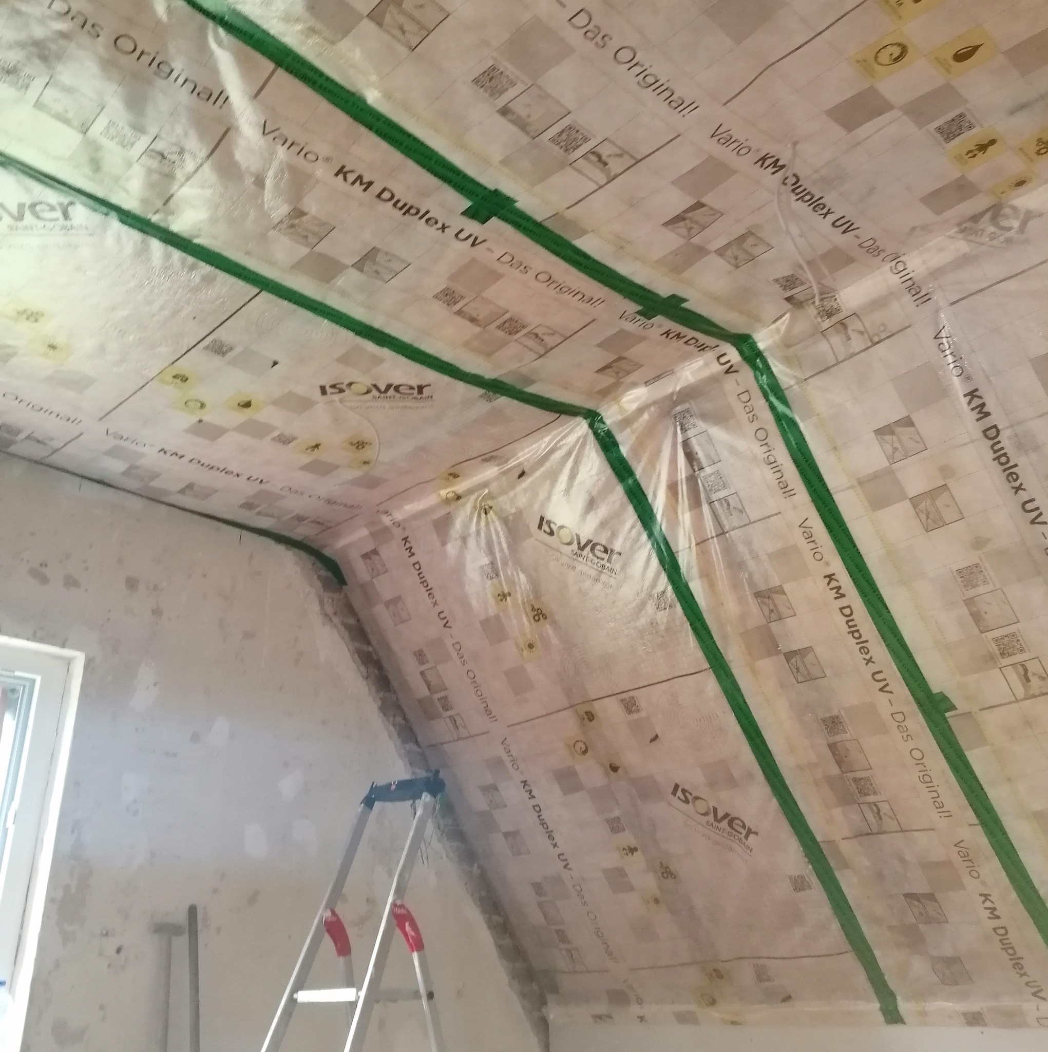 Poddasze w trakcie ocieplania, widoczna folia paroizolacyjna Isover Vario KM Duplex UV z zielonymi taśmami uszczelniającymi na krokwiach, drabina oparta o ścianę, surowy stan ścian.